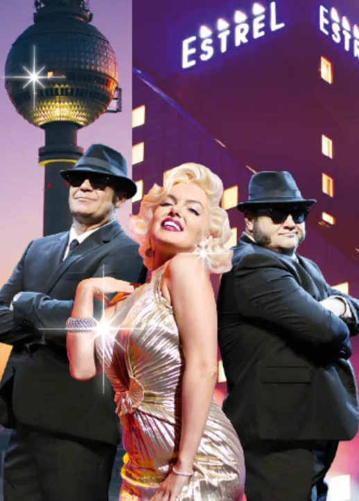 Drei Personen vor dem Hotel Estrel in Berlin, zwei Männer in schwarzen Anzügen und Hüten stehen mit verschränkten Armen, eine Frau in einem glänzenden, ärmellosen Kleid mit lockigen blonden Haaren posiert dazwischen.