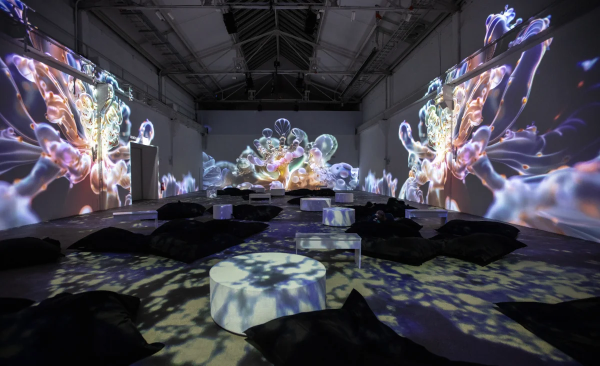 Großer Ausstellungsraum mit immersiven, organisch wirkenden Projektionen an Wänden und Boden sowie Sitzkissen und niedrigen Tischen im Raum