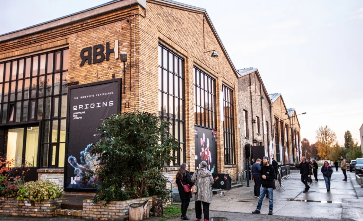 Backsteinfassade eines Industriegebäudes mit großen Fenstern und Plakaten zur Ausstellung »Origins – Ursprung des Lebens« vor dem Eingang, mehrere Personen stehen und gehen auf dem Gehweg davor