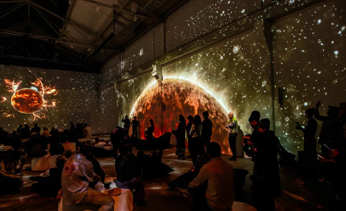 Menschen sitzen und stehen in einem dunklen Ausstellungsraum mit Projektionen von Sonne auf den Wänden im Rahmen der immersiven Ausstellung Origins – Ursprung des Lebens in Berlin