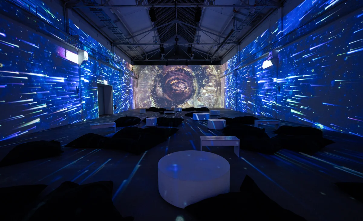 Große Gruppe sitzender und stehender Menschen in einem dunklen Ausstellungsraum mit immersiven, farbigen Projektionen an den Wänden, die ein spiralförmiges Muster an der Stirnwand zeigen
