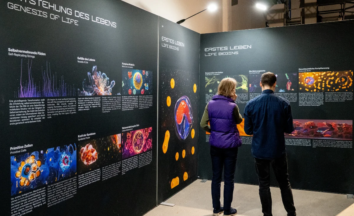 Zwei Personen betrachten eine Ausstellungstafel mit wissenschaftlichen Illustrationen und Texten zur Entstehung des Lebens in der Ausstellung "Origins – Ursprung des Lebens" in Berlin.