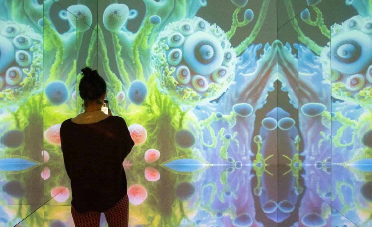 Person von hinten vor einer farbigen, organisch wirkenden Projektion mit kugeligen und röhrenförmigen Strukturen in einer Ausstellung