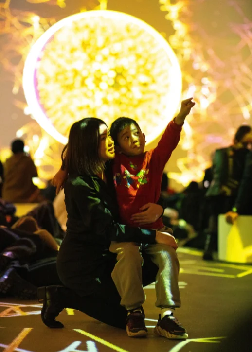 Eine Frau kniet auf dem Boden und hält ein Kind auf dem Schoß, das mit ausgestrecktem Arm auf eine große, leuchtende, gelbe Kugel im Hintergrund zeigt, umgeben von weiteren sitzenden Personen in einem dunklen Raum.