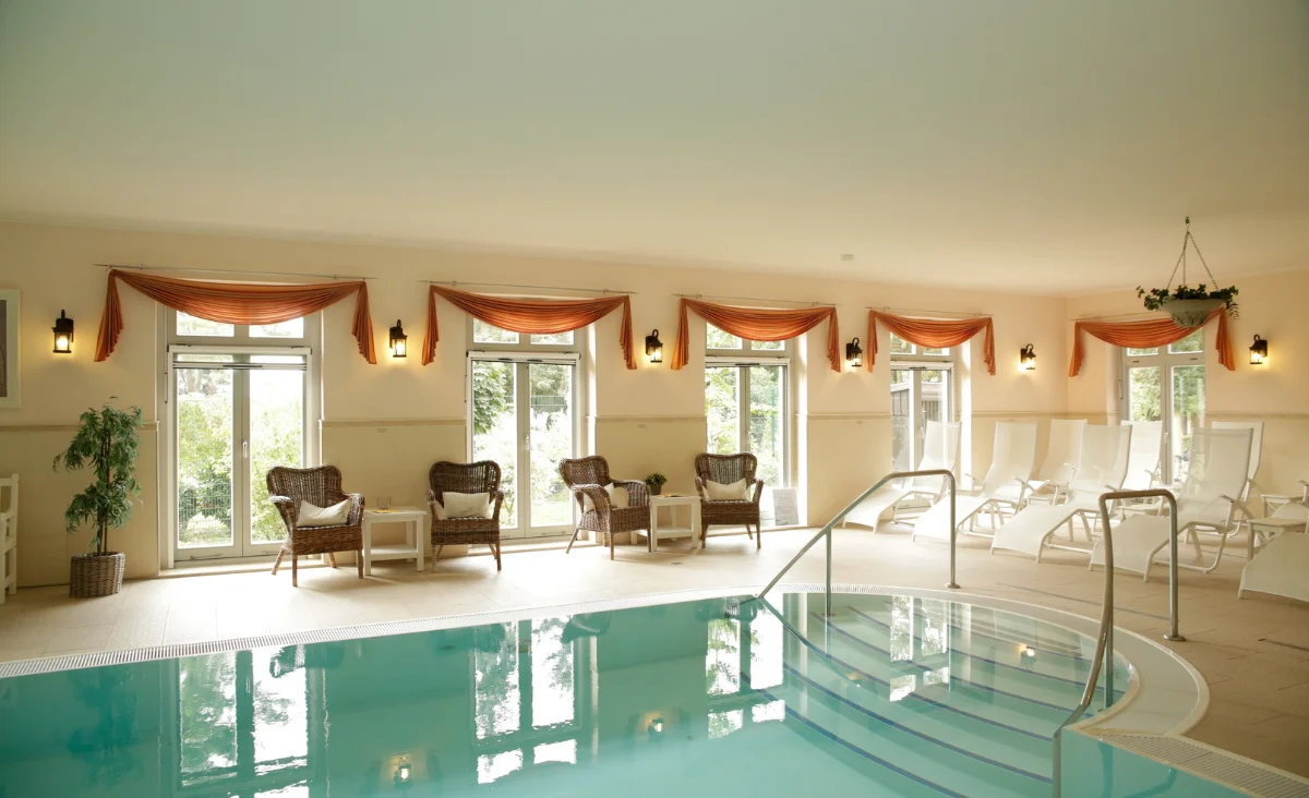 Innenraum eines Schwimmbads mit mehreren Fenstern, gepolsterten Korbsesseln, Pflanzen und Liegestühlen