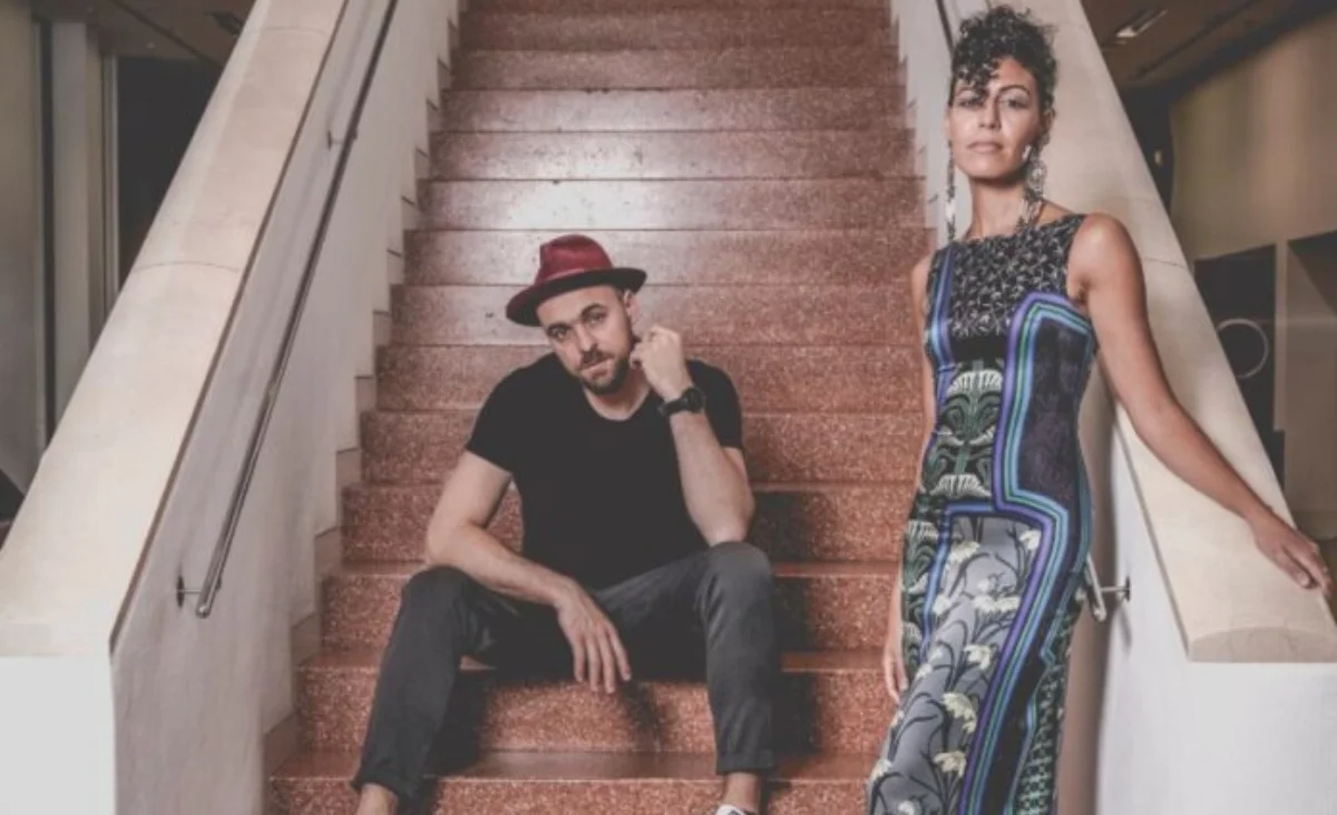 Ein Mann sitzt auf einer Treppe mit roten Stufen und trägt schwarze Kleidung und weiße Schuhe, eine Frau steht rechts neben der Treppe in einem langen, gemusterten Kleid