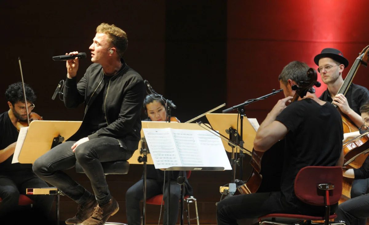 Mann sitzt auf Hocker und hält Mikrofon vor Mund, im Hintergrund Musiker mit Notenständern