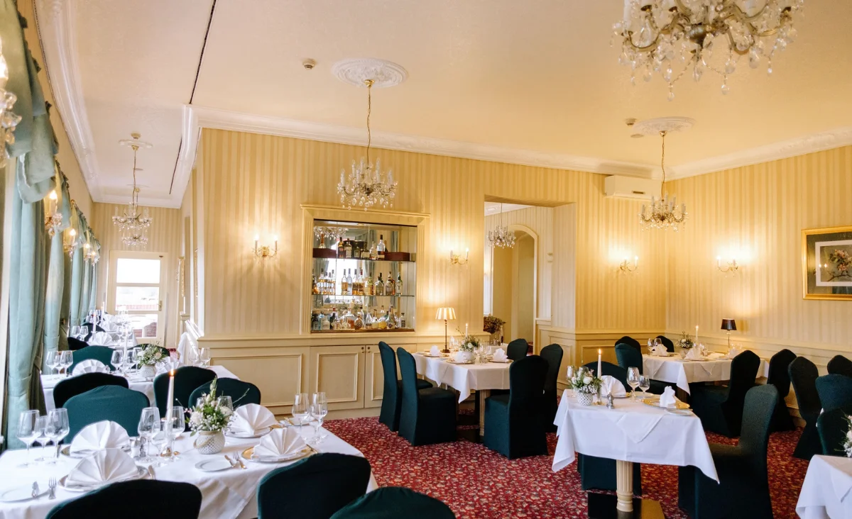 Eleganter Speisesaal mit mehreren gedeckten Tischen, grünen Stühlen, Kronleuchtern und einem Spiegel an der Wand