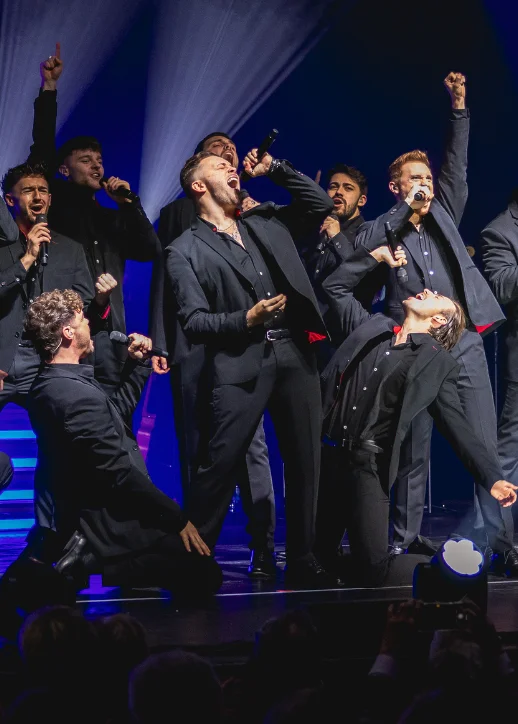 Mehrere Sänger in schwarzen Anzügen auf einer Bühne in dynamischen Posen mit erhobenen Armen und Mikrofonen: The 12 Tenors Konzert Berlin