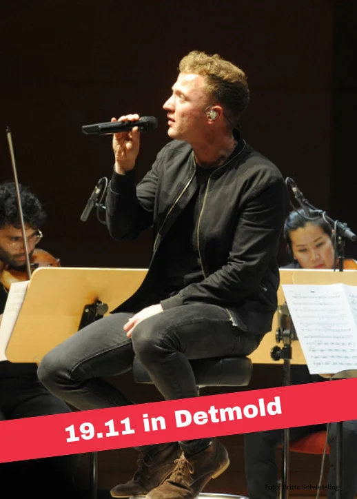 Mann sitzt auf Hocker und hält Mikrofon vor Mund, im Hintergrund Musiker mit Notenständern