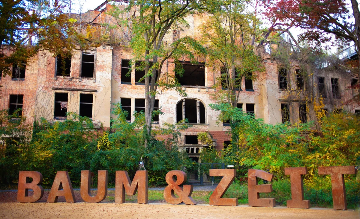 Große Metallbuchstaben mit der Aufschrift 'BAUM & ZEIT' vor einem verfallenen, mehrstöckigen Backsteingebäude, umgeben von Bäumen mit herbstlichem Laub.