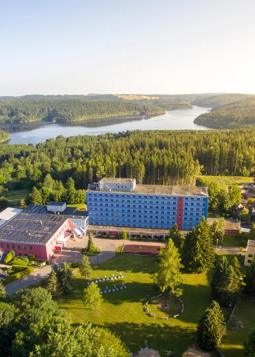 Das Hotel Blaues Wunder Eibenstock steht im Vordergrund einer Luftaufnahme. Das mehrstöckige Gebäude ist blau und hebt sich deutlich von den umliegenden, niedrigeren Gebäuden ab. Im Hintergrund erstreckt sich eine waldreiche Hügellandschaft mit einem großen, geschwungenen See. Die Umgebung ist von dichtem Wald und weitläufigen Grünflächen geprägt. Auf der Wiese vor dem Hotel sind mehrere Sitzgelegenheiten angeordnet. Die Aufnahme vermittelt einen Eindruck von ruhiger, naturnaher Lage.