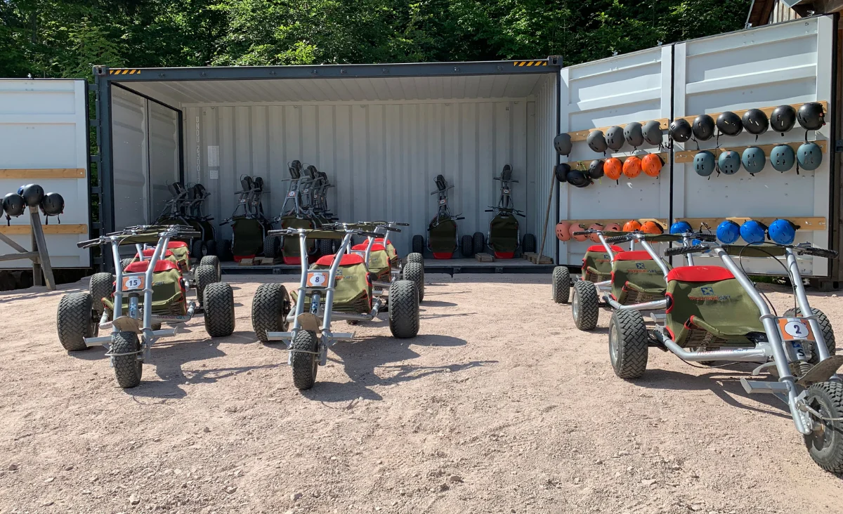 Mehrere geländegängige Dreiräder mit breiten Reifen stehen auf einem geschotterten Platz vor einem geöffneten Container. Im Container sind weitere Fahrzeuge desselben Typs abgestellt. An der rechten Containerwand hängen Schutzhelme in verschiedenen Farben auf Haken. Im Hintergrund ist dichter grüner Wald zu sehen. Die Szene gehört zu Wurzelrudis Erlebniswelt.
