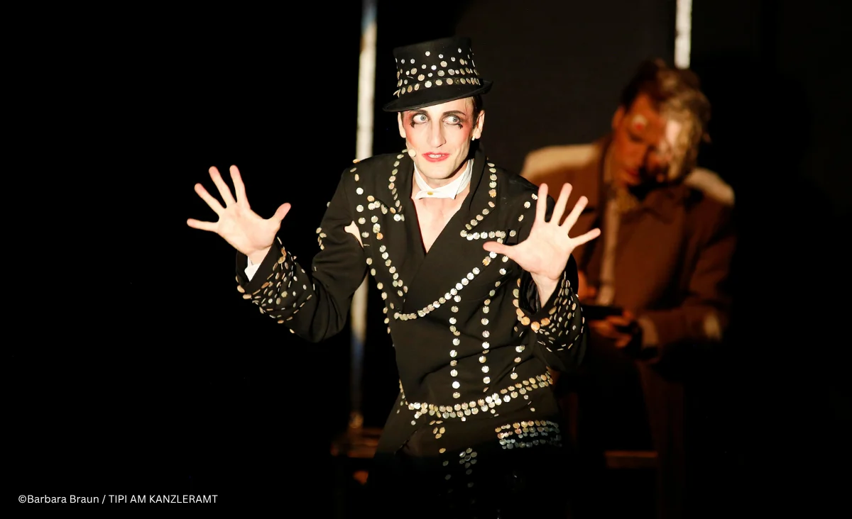 Ein Schauspieler in einem glitzernden Outfit führt eine dramatische Performance auf der Bühne auf, umgeben von dunklem Hintergrund.