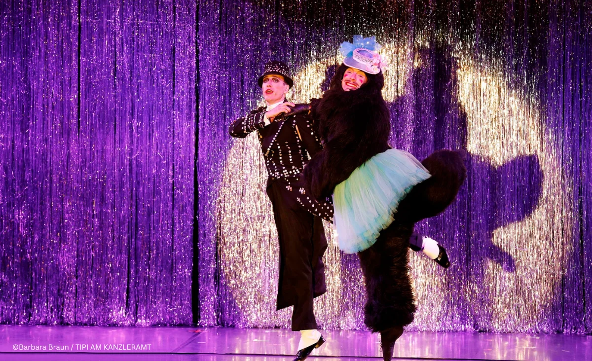 Zwei Personen stehen auf einer Bühne des Musicals »Cabaret Berlin! – das Berlin Musical« vor einem Vorhang aus silbernen Lamellen. Über ihnen leuchtet ein Schild mit der Aufschrift »KIT KAT KLUB« in großen, beleuchteten Buchstaben.