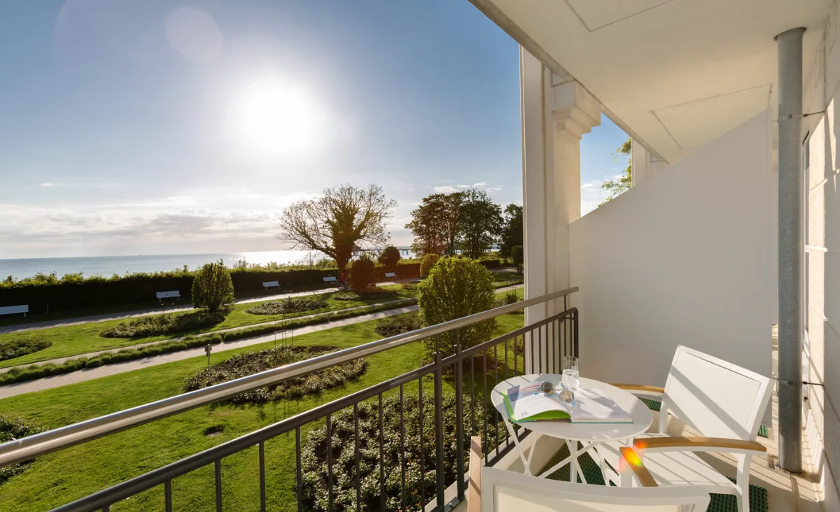 Balkon mit zwei weißen Stühlen und kleinem Tisch mit Glas und Buch, Blick auf Garten und Meer bei Sonnenschein