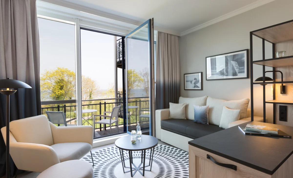 Helles Hotelzimmer mit Sofa, Sessel, rundem Tisch, offenem Balkonfenster und Blick auf Bäume aus Hotel Kaiserhof Heringsdorf
