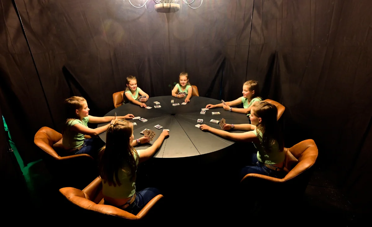 Sechs Kinder sitzen an einem runden schwarzen Tisch in einem dunklen Raum und spielen Karten