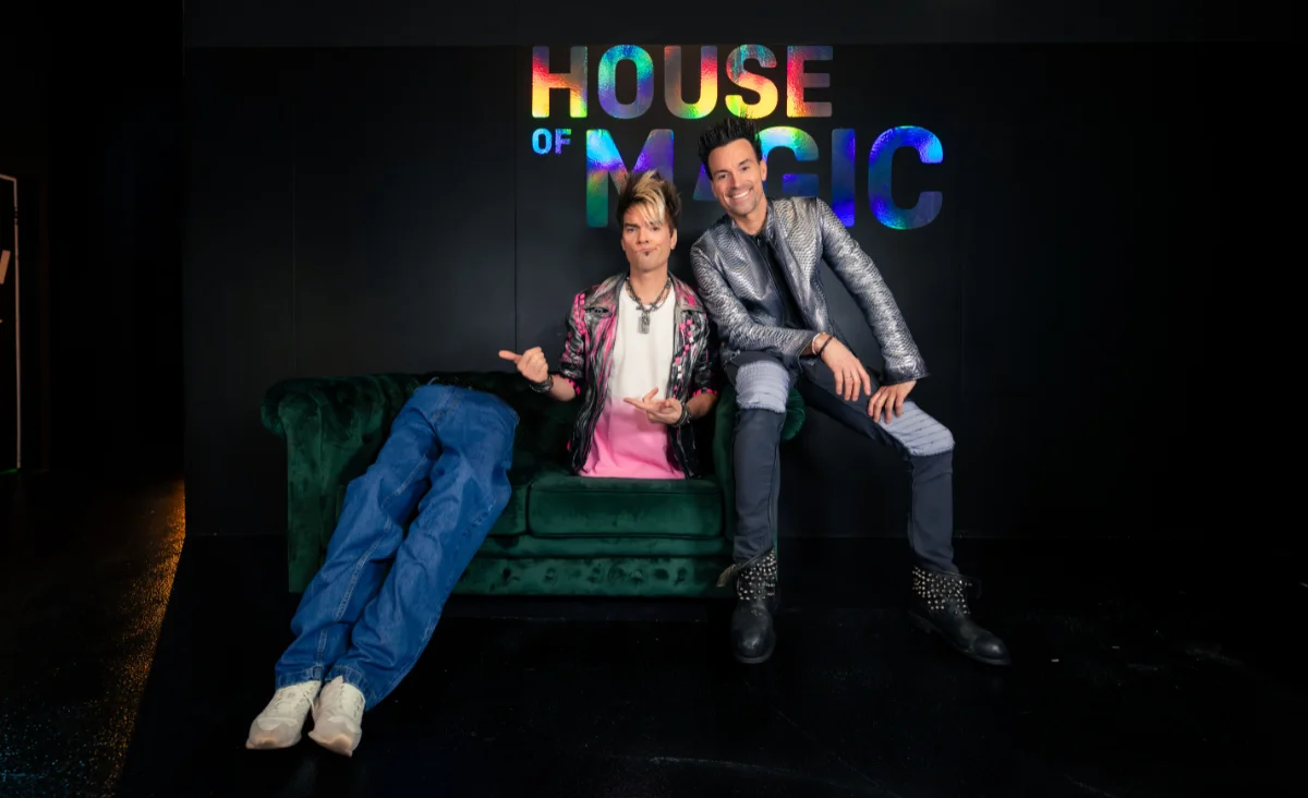 Zwei Männer in auffälligen Jacken posieren vor einer Wand mit dem Schriftzug 'HOUSE OF MAGIC', einer sitzt auf einem grünen Sofa, der andere lehnt sich daneben.