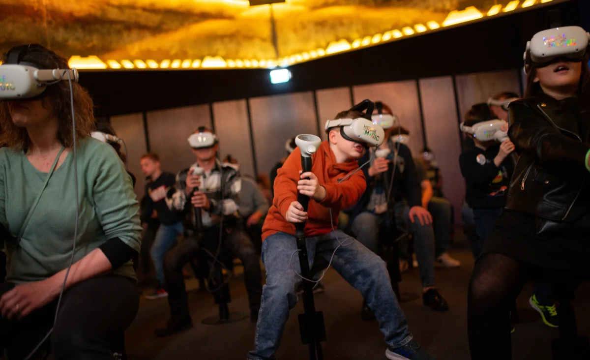 Mehrere Personen unterschiedlichen Alters sitzen in einem dunklen Raum und tragen VR-Brillen, ein Junge in der Mitte hält einen weißen Controller und lehnt sich nach vorne.