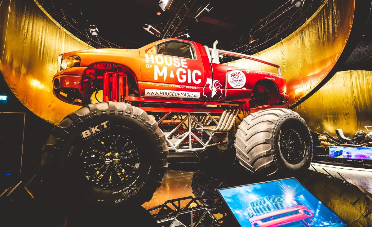 Roter Monstertruck mit großen Reifen und der Aufschrift 'HOUSE OF MAGIC' auf der Tür, ausgestellt in einer Halle mit goldfarbenem Hintergrund