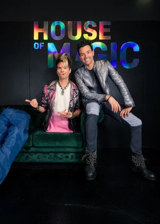 Zwei Männer in auffälligen Jacken posieren vor einer Wand mit dem Schriftzug 'HOUSE OF MAGIC', einer sitzt auf einem grünen Sofa, der andere lehnt sich daneben.