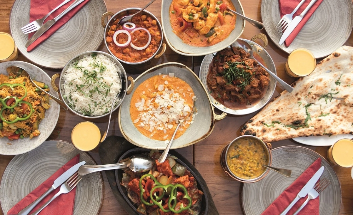 Tisch mit mehreren Schalen indischer Gerichte, Naan-Brot, Gläsern mit Getränken und Besteck auf grauen Tellern mit roten Servietten. Mit dem Wertgutschein Amrit Restaurants erleben.