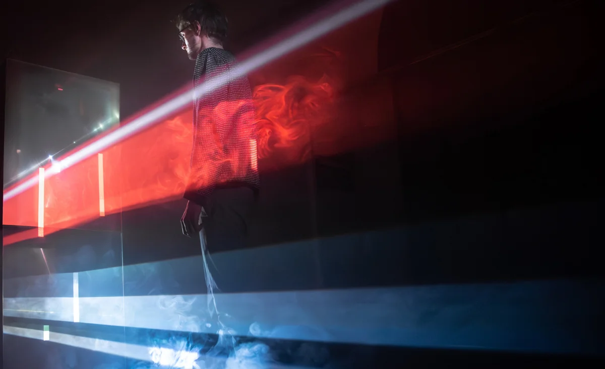 Person steht in einer dunklen Umgebung mit roten und blauen Lichtstreifen, die durch Nebel oder Rauch verlaufen